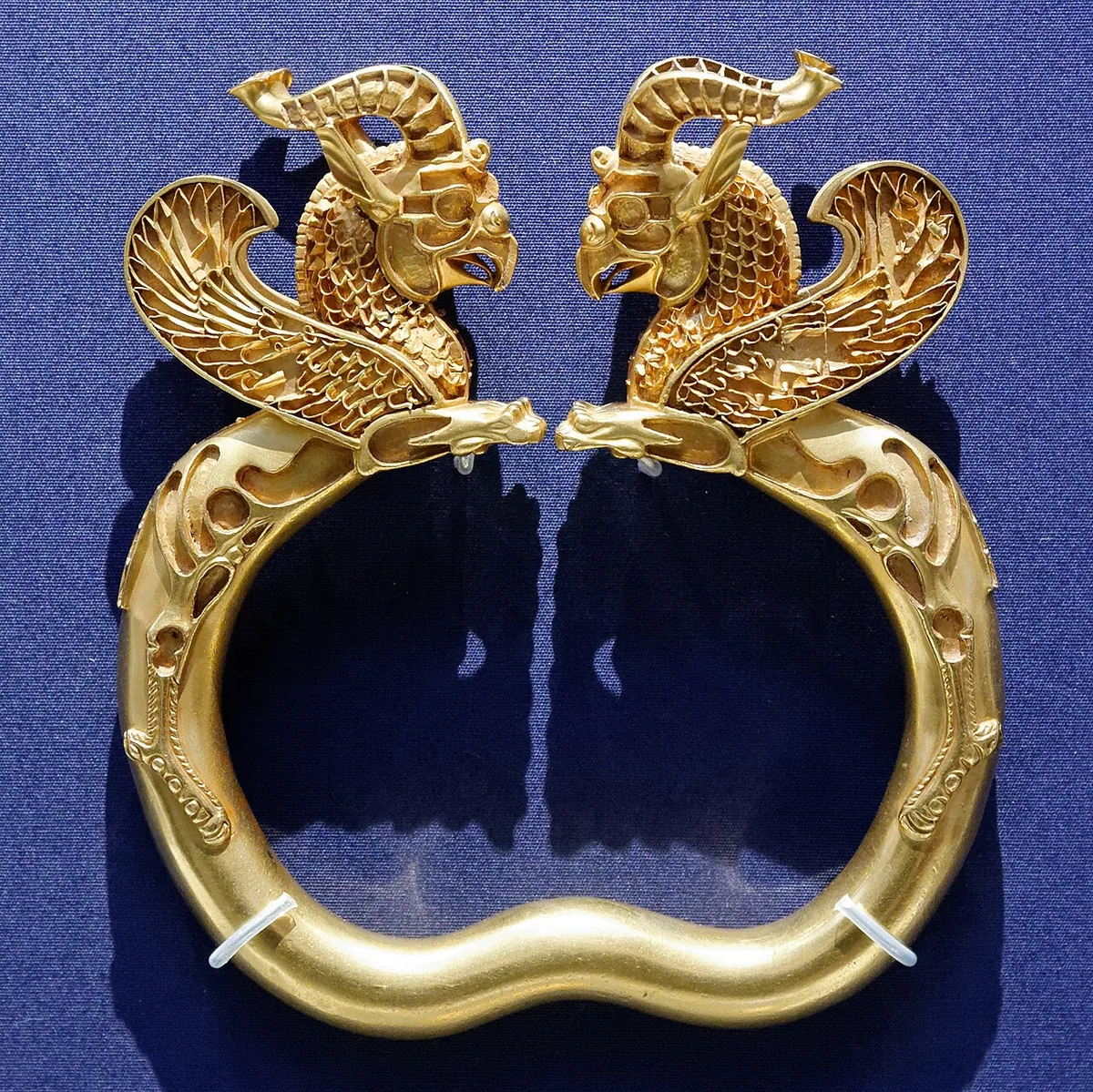 Gold armlet ending in two winged griffin heads facing each other, shown against a dark blue background